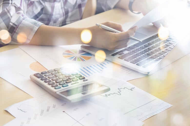 person sitting at a desk with a laptop, calculator, and papers with graphs on them