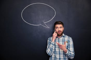 Happy handsome young man with beard talking on mobile phone standing over chalkboard background with blank speech bubble