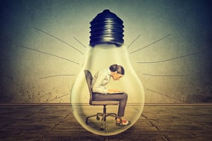 Woman working on laptop computer sitting inside electric light bulb isolated on gray office wall background