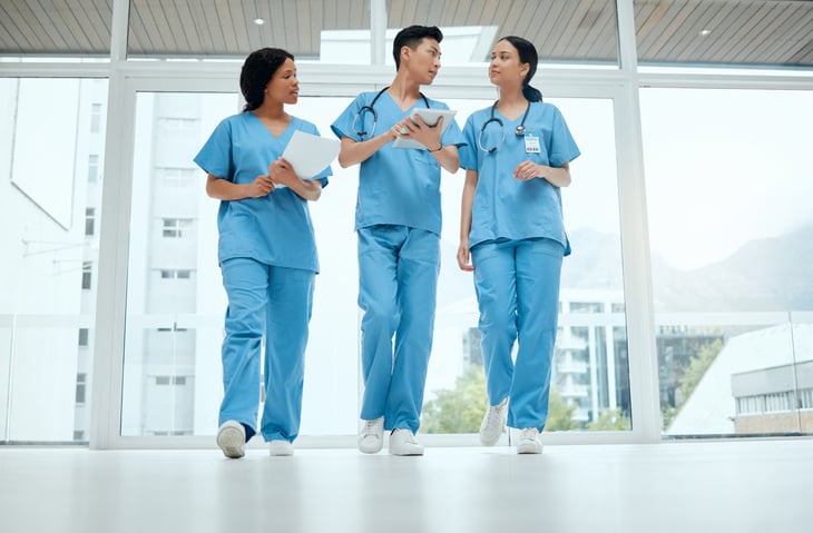 three nurses wearing scrubs walking down a bright hallway