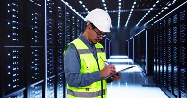 Engineer in high-visibility jacket and hard hat taking notes on a clipboard inside a data center.